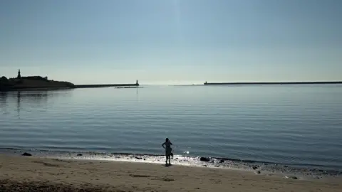 BBC Weather Watchers/ Deborah Johnson A woman standing on a beach looking out to the sea. Two piers can be seen in the distance.