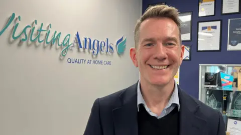 A man with short brown hair, wearing a blue suit and dark shirt and tie stands in front of a green-blue logo saying "Visiting Angels"