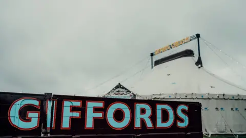 Giffords Circus A white circus tent is seen behind a sign reading "Giffords". The lettering is bright blue and red on a dark background.