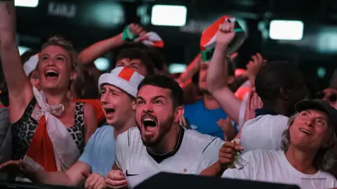 A fan in a white shirt and St George's Cross headband screams with clenched fists at Boxpark Croydon. Other fans around him shout and wave flags.