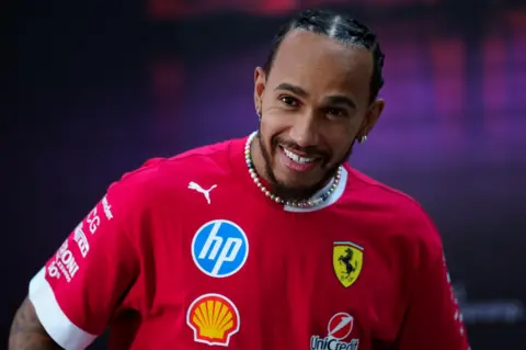 PA Media Lewis Hamilton in a Ferrari branded red shirt, with various other sponsors and branding on the t-shirt. He is smiling and wearing a beaded necklace.