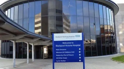 The outside of Blackpool Victoria Hospital showing a large round glass-fronted entrance beside a covered walkway and a blue sign saying 'welcome to Blackpool Victoria Hospital'