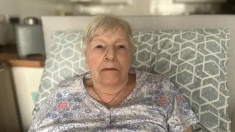 Catherine Nesbitt. She has short white hair, wearing a necklace and floral night gown, and lying in bed.
