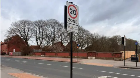 A stretch of road over a drain. Either side of the road are signs informing drivers they are entering a 20mph zone.