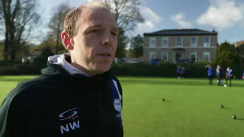A man stands on a bowling green, wearing a black hooded top. He has short light brown hair. Blurred out people playing bowls can be seen behind him.