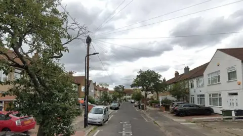A typical nondescript suburban street with houses on both sides of the road and various cars parked on the street and in front of the houses. 