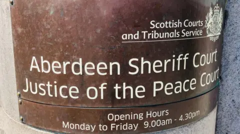 BBC Aberdeen Sheriff Court sign, bronze coloured, with white writing, says Scottish Courts and Tribunals Office, Aberdeen Sheriff Court Justice of the Peace Court, opening hours Monday to Friday 9am to 4.30pm.