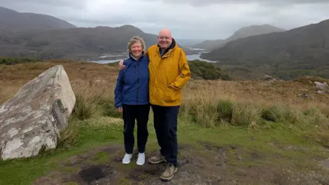 Older woman with grey hair in blue jacket and an older man in a yellow jacket smiling together with a rural backdrop including a lake and hills.