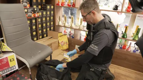 Trading Standards South West A Trading Standards officers wearing black and grey clothing and blue latex gloves rummages through a black bag in a shop to search for illegal tobacco products