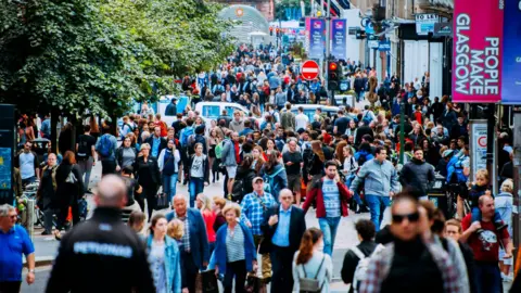 A picture of a crowded street in central Glasgow, with hundreds of pedestrians walking both towards and away from the camera. Green trees are visible on the left of frame, the entrance to a subway station is at the far end of the street in the middle, and a "People Make Glasgow" banner adorns a lamp post to the right.