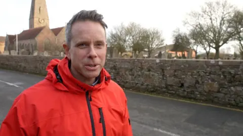 Man wearing bright red coat stood near road with a church behind. He has brown hair