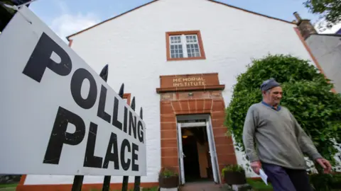 Getty Images A man wearing a grey jumper and a blue hat leaves after casting his vote at the Moniaive polling station in Dumfries and Galloway. The building has white walls and the doorway is made of red sandstone. A sign on the left of the image says Polling Place.