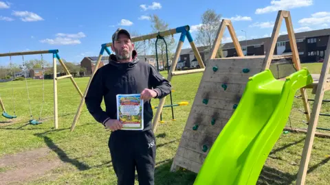 Jonny Humphries/BBC Lewellyn Mitchell, 45, who has a grey beard and is wearing a green baseball cap with the peak turned backwards and a black Duffer branded hoodie, looks at the camera in front of wooden children's play equipment on a grassy field, surrounded by terraced houses. He is holding a stack of 'park activity waivers' on A4 paper printed with a colourful cartoon image of children playing.
