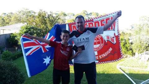 Allan McCarlie Jamie and Allan in 2017 before heading to Scotland for the cup final. Jamie is holding an Australia flag, which is a blue background with white stars, and a red, white and blue Union Jack style flag in the corner. He is wearing a red Aberdeen FC jersey. Allan is holding a red flag which says "Aberdeen" and has a blue and white saltire cross on one side. He is wearing a white and black Aberdeen jersey. They are standing in a garden.