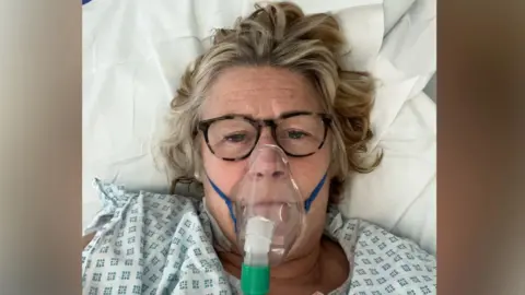 Rachael Coyne A woman in her 50s is lying in a hospital bed with an oxygen mask on her face. She looks very poorly and is wearing a surgical gown.