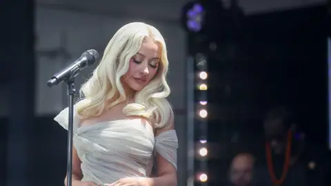 Getty Image Christina Aguilera standing on stage next to a microphone. She is looking down with her eyes closed. She has long blonde hair and is wearing a cream dress that appears to be sparkling. 