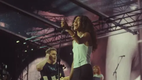 Polydor Label Group Sam Fender appears in the background holding onto his ear, wearing a black T-Shirt with his guitar also visible. Olivia Dean is at the front singing into a microphone wearing a white tank top.