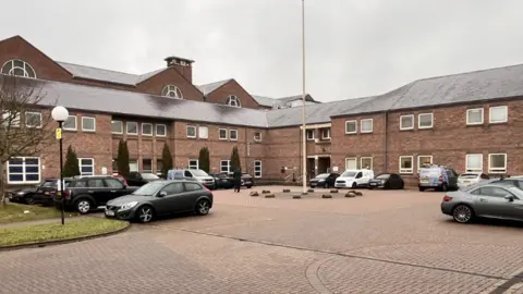 Norwich Crown Court forecourt with a brickweave car parking area dotted with parked cars and an L-Shaped, two-storey brick building in the background 