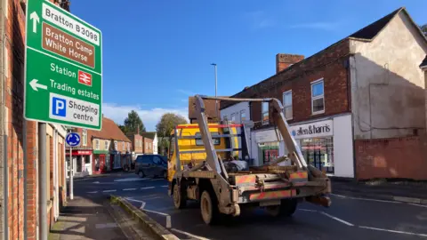 The road going up to a mini roundabout in the middle of Westbury with two-storey red brick buildings and homes on either side, with a skip lorry coming up to the roundabout and a road sign on the left