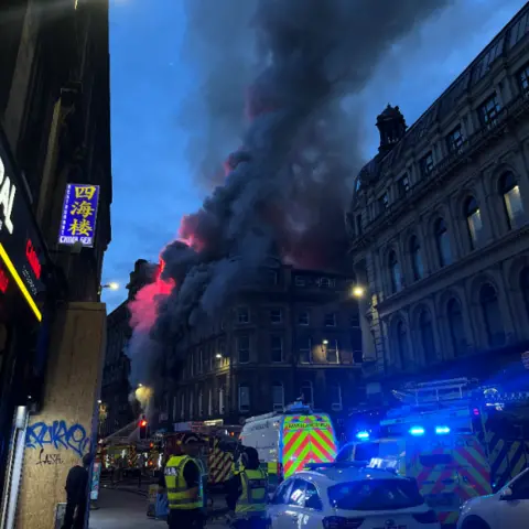 PA Media Huge thick clouds of black smoke billow into the evening sky with several emergency service vehicles parked in the street