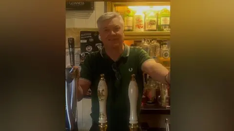 Connor Devine A man with grey hair and wearing a polo top is stood behind a bar with beer pumps in front of him. He is wearing a green top and drinks can be seen on shelves behind him.