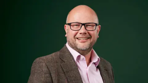 Prologis A bald man with a beard is smiling at the camera, he is wearing black glasses. He is wearing a brown tweed jacket and a pink shirt