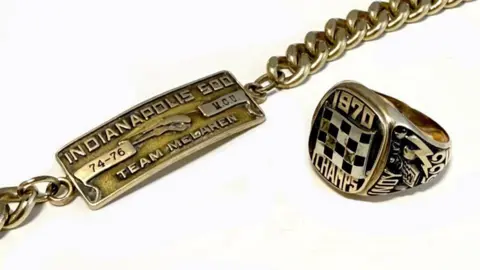 A gold and black signet ring with the words "1970 CHAMPS" on the front and a gold bracelet with the words "INDIANAPOLIS 500 74-76 TEAM MCLAREN"