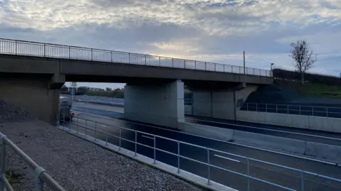 A concrete bridge with metal railings over a road showing two sides of the carriageway.