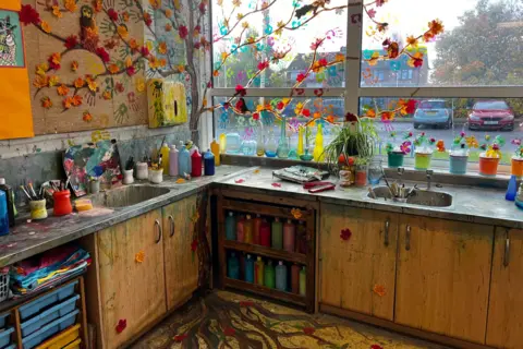 Josh Parry / BBC The corner of an art classroom where Heartstopper was filmed. The countertops and sinks are covered with paint bottles, and on the wall and window is a mural of a tree with fall leaves, its roots painted on the floor