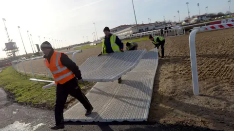 PA Media Workers in hi-vis vests carrying large floor mats at the racecourse. They are crossing the sandy race track.