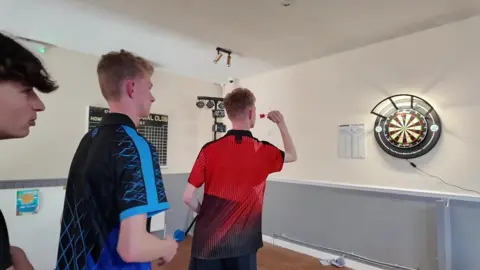 Twins Harry and Alfie playing darts. Alfie is holding a dart in front of a dart board. Harry is standing behind.