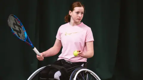 Barbara Leatham for LTA A girl in a wheelchair sits side-on while holding a tennis racket in one hand and a tennis ball in the other. She is wearing a pink T-shirt and black trousers, and has auburn hair tied back.
