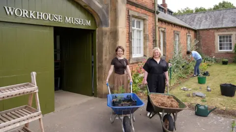 Museum director Alexa Vernon Museum and curator Dr Laura Allan stand with wheelbarrows outside the entrance to a workhouse museum.