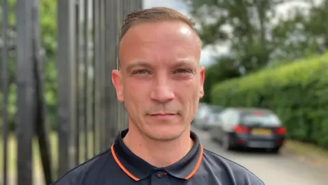 Freddie Cooper is looking directly into the camera, he is wearing a black and orange top. He's standing outside the gates of Saffron Hill cemetery in Leicester.