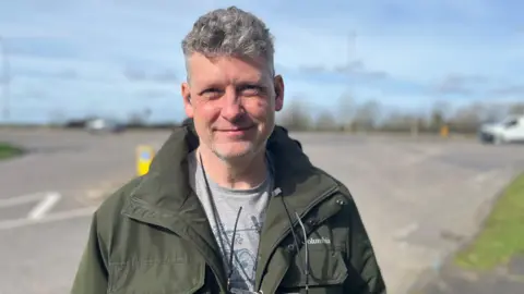 David May looking into camera with the A10 junction behind him. David has grey hair, is wearing a dark green jacked and grey top, with glasses on a string hanging from his neck.