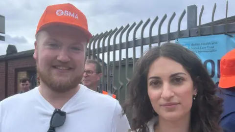 Adam McConnell is wearing an orange BMA hat, a white t-shirt and has hung his sunglasses on it. He has a ginger beard. Kiana has brown hair and is wearing a white jacket.