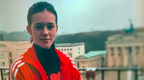 Brunette white teenage girl standing on a balcony. Background is blurred buildings. She is wearing a red sports jacket and black polo neck top.