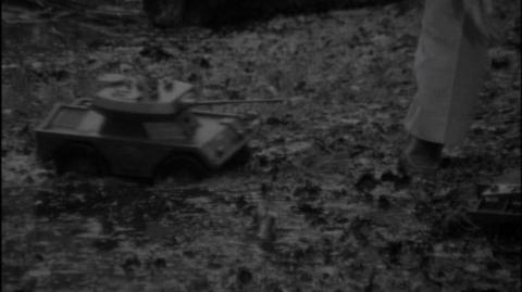 Black and white. Model tank on the ground in forest next to a leg, shoe and trouser leg vision.