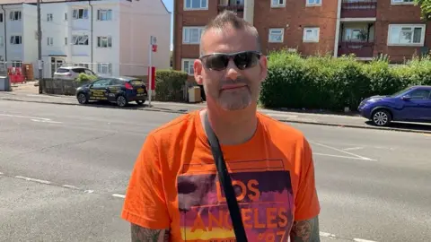 Picture shows a white man with short brown hair and sunglasses, with a bit of stubble wearing a bright orange t shirt that says 'Los Angeles'. He is wearing sunglasses and has tattoos on his arms. He is standing at the side of a street and behind him is a road and some flats.