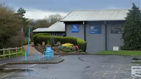 Google Street View of Two Riversmeet leisure centre viewed from the car park on a grey rainy day. The grey warehouse-style building has a pitched roof. A curved hedge and a rockery area planted with shrubs separates the car park from the entrance.