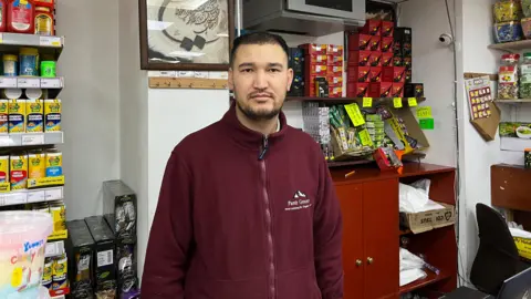 George Carden/BBC Ishak Karimi in a dark red fleece reading Pamir Grocery behind the counter in his shop.