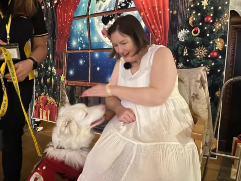 Nicola Haseler/BBC Katie sits on a chair and leans over to pat Ralph on the head - he is sitting next to her and looking up at her. She wears a white dress and has a flower in her hair and looks cheerful. Helen is standing next to Ralph and holding his lead.