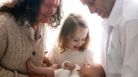 Aga Kowlska Photography shows a family picture that shows a mummy wearing a porridge cardigan and white shirt with a child in a white baby, a girl in a white top and a man in a white shirt