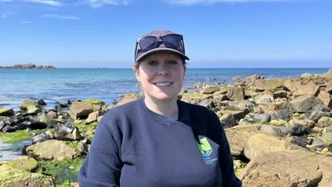 A woman wearing a cap and sunglasses standing in front of the sea and rocks
