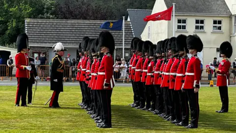 The governor, if full ceremonial dress, inspects two rows of the Grenadier Guards, who are wearing red tunics, black trousers and black bearskin caps.