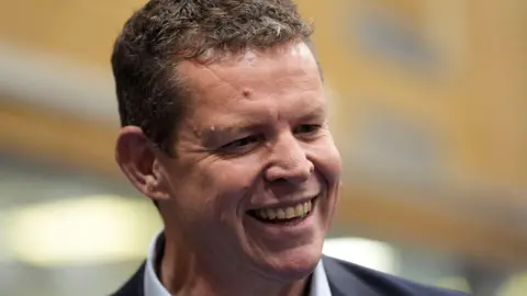 PA Media Rhun ap Iorwerth tersenyum dengan hanya kepala dan bahunya yang terangkat. Dia mengenakan jas gelap dan kemeja putih.