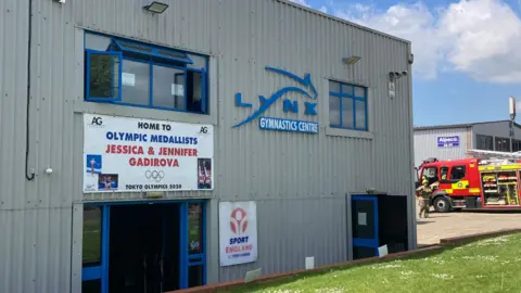 Alex Pope/BBC The outside of a gym, with banners on it and a fire engine to the right. The building is grey, with blue trim.
