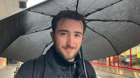 Vikki Irwin/BBC A man is standing on the street under a black umbrella, he is wearing a grey tweed coat and is carrying a back pack.