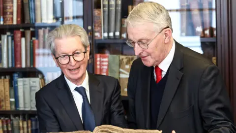 Two men wearing glasses and navy suits with white shirts and ties look at old copies of a newspaper. The copies have been bound together in a large book. A shelf with books is behind them.