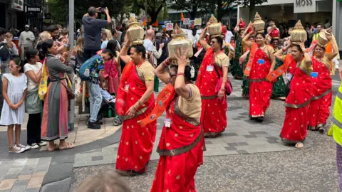 A group of more than six women in red and beige saris hold up golden-coloured vessels on their heads as they walk through a street past spectators, who are watching or taking pictures.